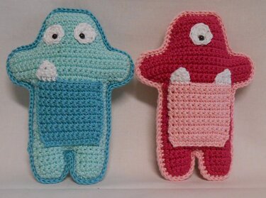 Zwei gehäkelte Monsterfiguren, eine türkis mit zwei Augen und eine pink mit einem Auge, beide mit kontrastfarbener Bauchtasche und weißen Zähnen.