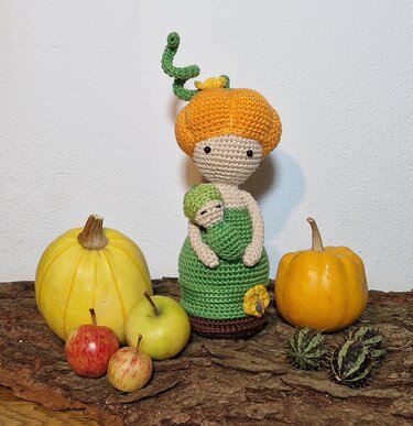 Gehäkelte Amigurumi-Puppe mit orangefarbenem Kürbiskopf, grünem Kleid und kleinem Baby in den Armen, arrangiert zwischen Kürbissen und Äpfeln auf Baumrinde