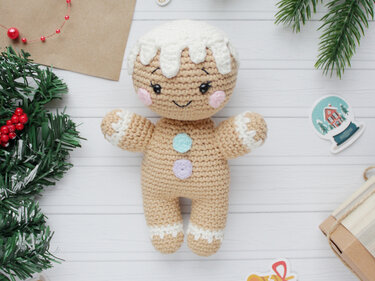 Gingerbread amigurumi doll with white icing trim, pastel buttons and a stitched smile on white wooden boards with evergreen decorations