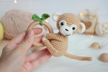 Hand holding a wooden teething ring with a beige crocheted monkey attached, yarn balls blurred behind