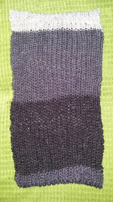 Gestrickter Loop-Schal in Grautönen mit sichtbarer Maschenstruktur auf hellem grünem Untergrund.