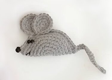 Grau gehäkelte Amigurumi-Maus mit großen Ohren, Perlenaugen, schwarzer Nase und gedrehtem Schwanz auf hellem Hintergrund