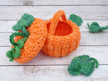 Gehäkelter orangefarbener Kürbiskorb mit Deckel, Henkel und grünen Blättern auf heller Holzfläche