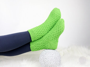 Pair of lime-green crocheted ankle socks worn over navy leggings, feet resting on a patterned grey ball on a white faux-fur rug.