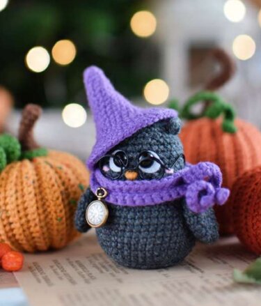 Detailaufnahme einer grauen gehäkelten Eule mit lila Hut, Schal, Brille und Taschenuhr, vor gehäkelten Kürbissen.