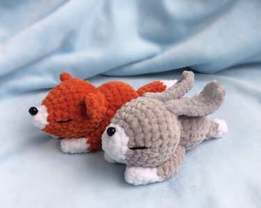 Oranger Amigurumi-Fuchs und grauer Amigurumi-Hase liegen nebeneinander auf hellblauem Stoff.