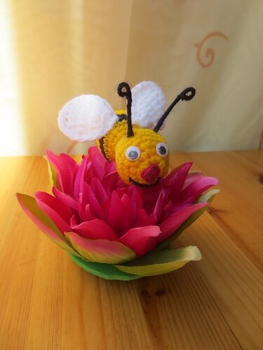 Gehäkelte Amigurumi-Biene mit Wackelaugen, weißen Flügeln und schwarzen Fühlern in einer pinken Blüte auf einem Holztisch