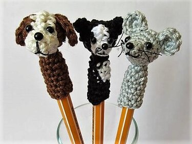Drei gehäkelte Tieraufsätze auf Bleistiften: braun-weißer Hund, schwarz-weißer Hund und graue Maus.