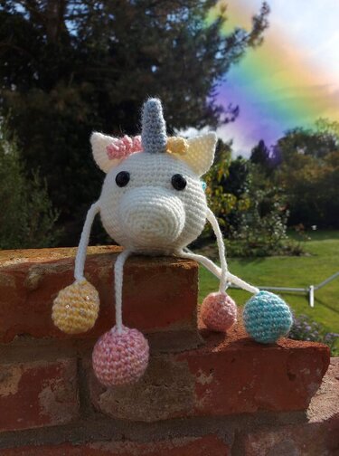 Weißes gehäkeltes Einhorn-Amigurumi mit Horn und vier pastellfarbenen Kugelbeinen auf Ziegelmauer vor Garten und Regenbogen