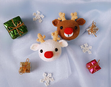 Two crocheted amigurumi reindeer head ornaments, one white and one brown, with red noses and small antlers, surrounded by tiny wrapped gifts and crochet snowflakes on pale blue fabric.