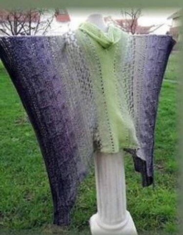 Gehäkelter Poncho mit Rollkragen und Farbverlauf in Grün, Grau und Lila auf einer weißen Säule im Freien.