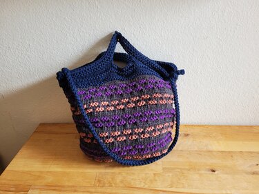 Handgestrickte Tasche in Dunkelblau, Grau, Lila und Pfirsich mit horizontalem geometrischem Muster auf Holzoberfläche.
