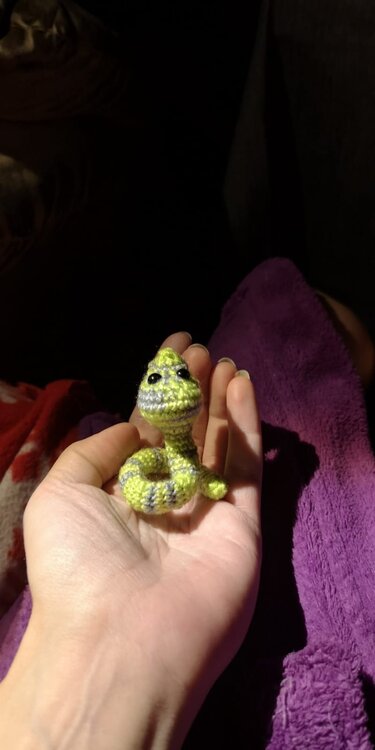 Winzige gehäkelte Schlange in Grün und Grau mit geringeltem Körper, gehalten in einer Hand.