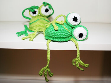 Two green crocheted amigurumi frogs with large white-and-black eyes, one hanging legs off a white shelf