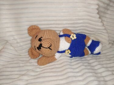 Nahaufnahme eines gehäkelten Teddybären in blauer Latzhose mit Blumenknöpfen und gestreiften Gliedmaßen auf weißer Decke.