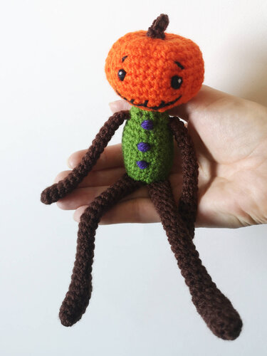 Hand hält gehäkeltes Kürbis-Amigurumi mit grünem Körper und langen braunen Beinen