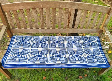 Blue and white crocheted blanket with repeating geometric square pattern on a rustic wooden bench.