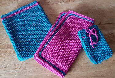Zwei gestrickte Tücher in Pink, ein türkisfarbenes Tuch und ein kleiner türkisfarbener Beutel mit pinkem Band auf Holz.