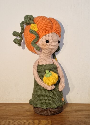 Gehäkelte Amigurumi-Puppe mit orangefarbenem Kürbiskopf, grünem Kleid und kleinem gelben Kürbis.