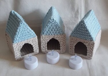 Drei gehäkelte Miniaturhäuser in Blau und Beige mit LED‑Teelichtern davor.