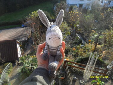 Handgehaltener, gehäkelter Amigurumi-Esel in Grau und Weiß vor unscharfem Landschaftshintergrund.