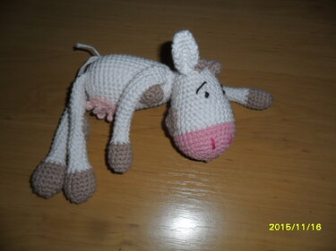 Gehäkelte Amigurumi-Kuh mit weißem Körper, braunen Flecken und rosa Schnauze liegend auf Holzboden
