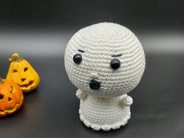 Weißer gehäkelter Amigurumi-Geist mit schwarzen Augen, daneben zwei unscharfe Halloween-Kürbisse auf dunklem Hintergrund.