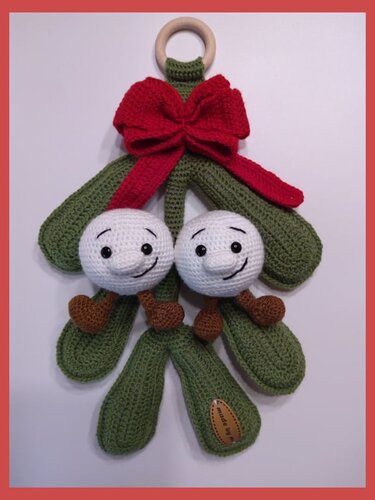 Gehäkelter Mistelzweig mit zwei weißen Amigurumi‑Kugeln, roter Schleife und Holzring zum Aufhängen.