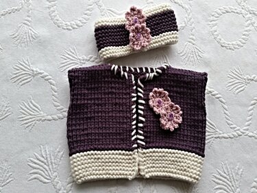 Gestrickte ärmellose Babyweste und Stirnband in Lila und Creme mit rosa Blumenapplikationen.