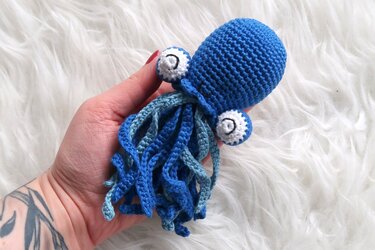 Hand hält blaues gehäkeltes Amigurumi mit gedrehten Tentakeln auf weißem Fell