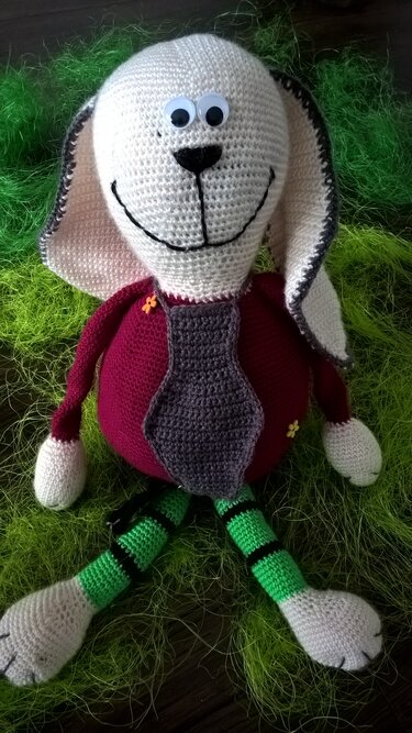 Gehäkelter Amigurumi-Hase mit weißem Kopf, langen Ohren, dunkelrotem Körper, grauer Krawatte, gestreiften Beinen und Wackelaugen auf grünem Dekogras.