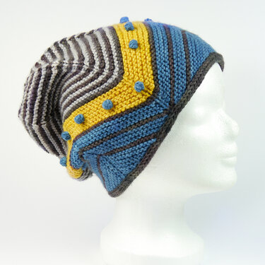 Slouchy knitted beanie with blue, yellow, and grey stripes and blue bobbles on a white mannequin head, viewed from the side.