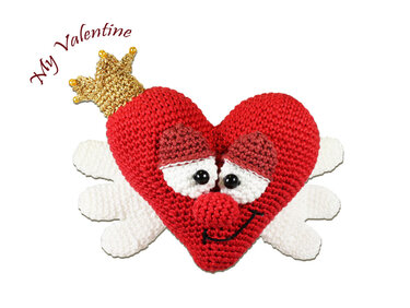 Rotes gehäkeltes Amigurumi-Herz mit Gesicht, weißen Flügeln und goldfarbener Krone auf weißem Hintergrund.
