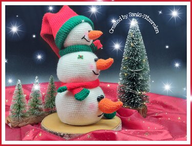 Gehäkelte gestapelte Schneemänner mit roten Mützen, grünen Schals und orangen Nasen auf rotem Stoff mit kleinen Tannen