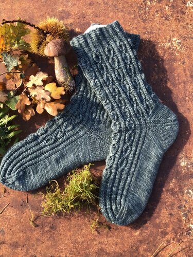 Gestrickte blaugrüne Damensocken mit Zopf- und Rippenmuster auf rostigem Untergrund, dekoriert mit Moos, Eichenblättern und Kastanienhülle