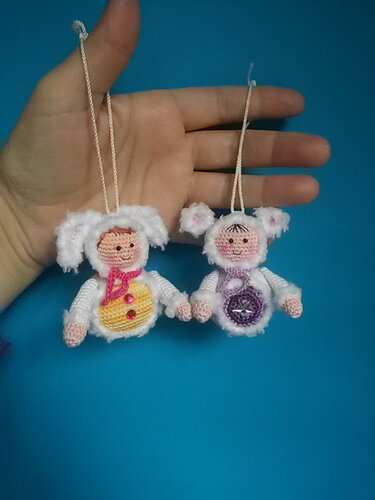 Zwei gehäkelte Amigurumi-Anhänger mit weißen flauschigen Kapuzen und Aufhängeschlaufen vor blauem Hintergrund, gehalten von einer Hand.