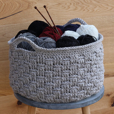 Gehäkelter grauer Korb mit Wollknäueln und Stricknadeln auf einem Hocker