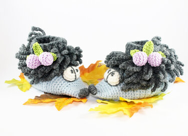 Paar gehäkelter Igel-Babyschuhe in Grau und Hellblau mit lila Beeren auf orange-gelben Herbstblättern.
