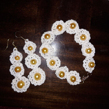 Weiße gehäkelte Blütenohrringe und passendes Armband mit goldfarbenen Perlen auf dunklem Holz.