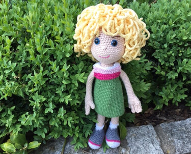 Gehäkelte Amigurumi‑Puppe mit lockigen blonden Haaren, grünem Kleid und grauen Schuhen vor Buschwerk.
