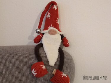 Gehäkelter Weihnachtswichtel mit roter Zipfelmütze, weißem Bart und Glocke, sitzend auf einer Sofalehne