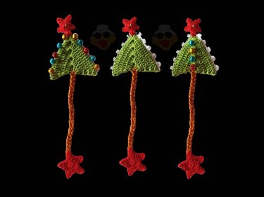 Drei gehäkelte Weihnachtsbaum-Lesezeichen mit roten Sternen, orangefarbener Kordel und bunten Glöckchen auf schwarzem Hintergrund.