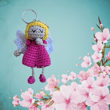 Gehäkelter Engel-Schlüsselanhänger mit rosa Kleid, gelbem Haar, goldfarbener Aureole, weißen Flügeln und Metallring vor aquamarinfarbenem Hintergrund mit Kirschblüten-Illustration.