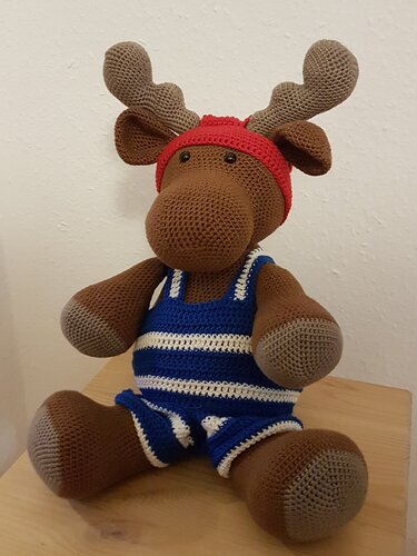 Gehäkelter Amigurumi-Elch mit rotem Stirnband und blau-weiß gestreiftem Overall, sitzend auf einem Holztisch