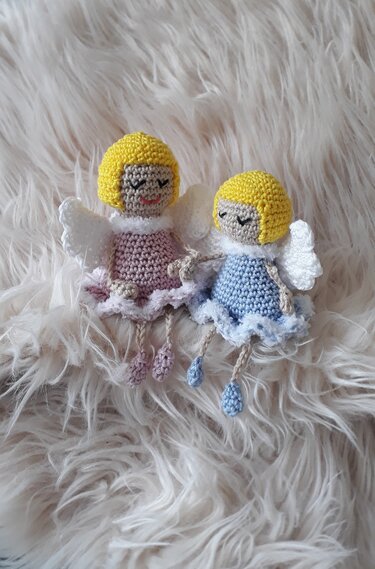 Zwei gehäkelte Amigurumi-Engelchen mit gelben Haaren, weißen Flügeln und Kleidern in Rosa und Hellblau auf flauschigem Untergrund.