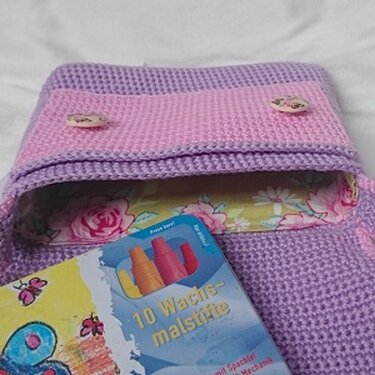 Gehäkelte lila-rosa Tasche mit Klappe, zwei Knöpfen, floralem Innenfutter und einer Packung Wachsmalstifte davor.