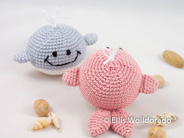 Zwei gehäkelte Amigurumi-Wale in Rosa und Hellblau mit gesticktem Gesicht auf weißem Hintergrund, umgeben von Muscheln.