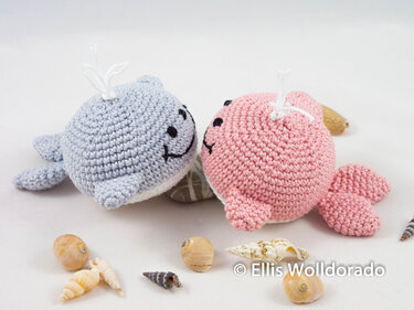 Zwei gehäkelte Amigurumi-Wale in Pastellblau und Rosa, liegend neben Muscheln