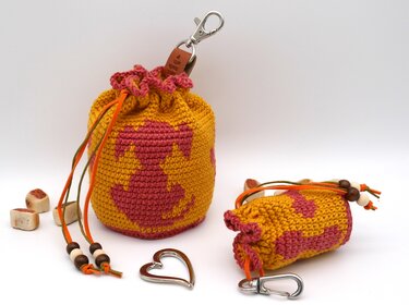 Zwei gehäkelte Leckerlibeutel in Senfgelb und Rosa mit Hundemotiven, Kordelzug, Holzperlen und Karabinerhaken auf weißem Hintergrund.