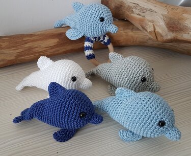 Fünf gehäkelte Amigurumi-Delfine in Blau, Weiß und Grau, einer davon an einem umhäkelten Holzring.